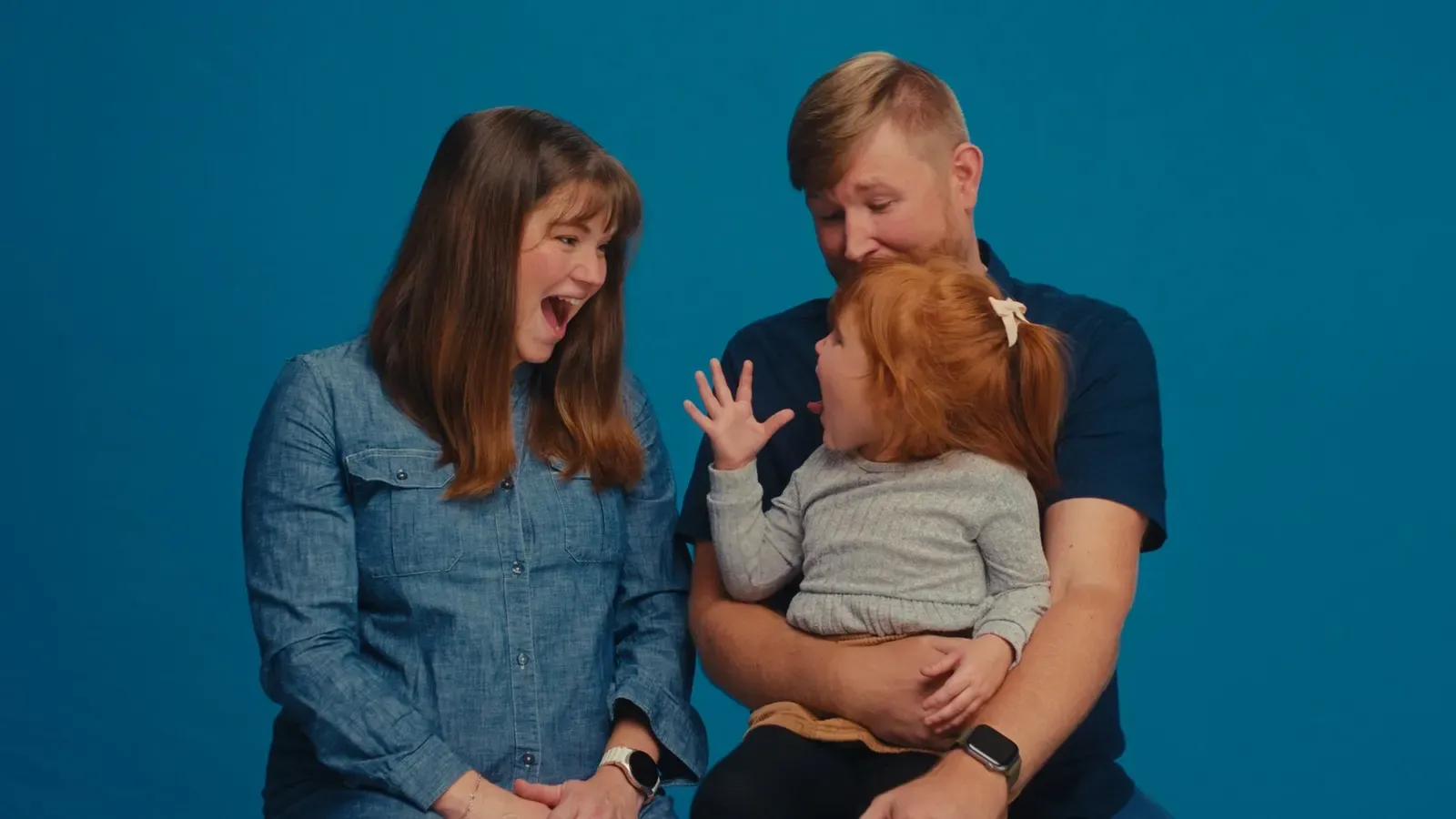 The Hartmans laughing together with their young daughter against a blue backdrop